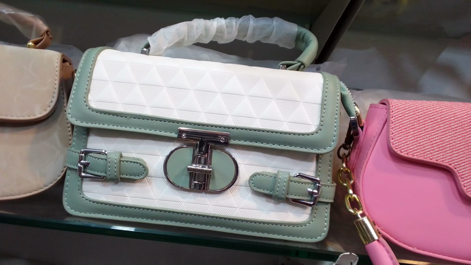 This chic handbag features a striking two-tone design with a white, diamond-quilted pattern on the front flap and body, framed by a soft, seafoam green trim. The bag is secured with a polished, silver-toned clasp and two decorative buckle straps. It has a sturdy top handle, wrapped for protection, and a detachable silver chain strap, offering versatile carrying options